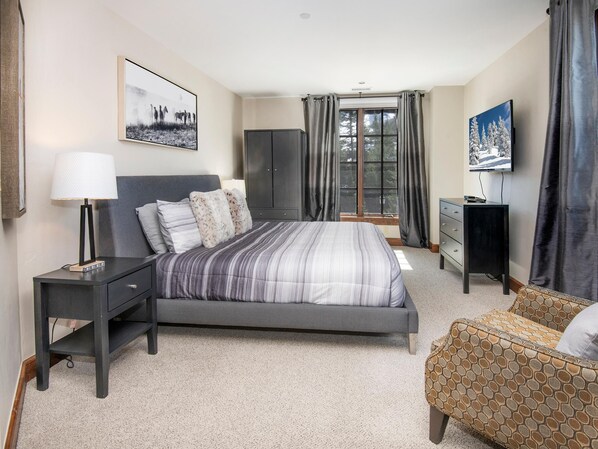 A bedroom with a bed, bedside table, lamp, wardrobe, TV, and a patterned armchair. - Ascent 301, 3BD Condo in Avon With Amenities (Avon)