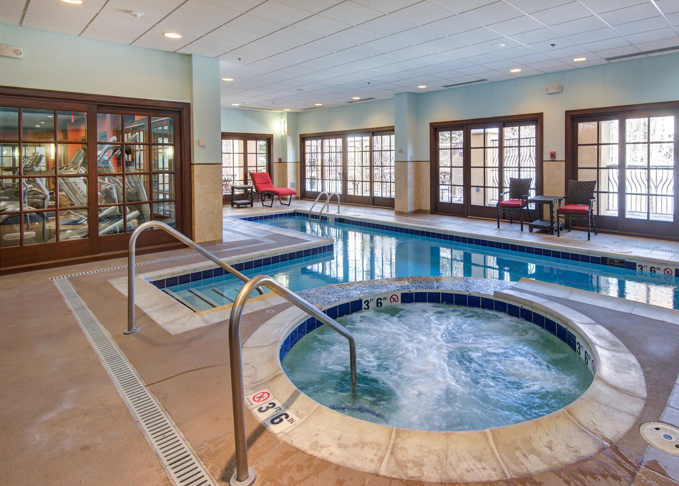 Piscina interior de hotel con jacuzzi, sillones de descanso y una zona cerrada con equipo de ejercicio.