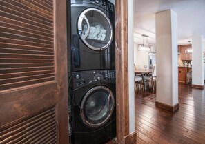 A modern laundry room with a stacked washer and dryer, wooden flooring, and a view into a dining area. - Ascent 301, 3BD Condo in Avon With Amenities (Avon)