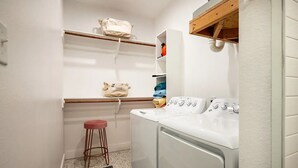 Un cuarto de lavado con lavadora, un taburete, repisas con bolsas y una alfombra estampada.