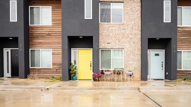 Un edificio de varios pisos con puertas numeradas, una puerta amarilla y un pequeño espacio al aire libre con sillas y plantas en macetas.