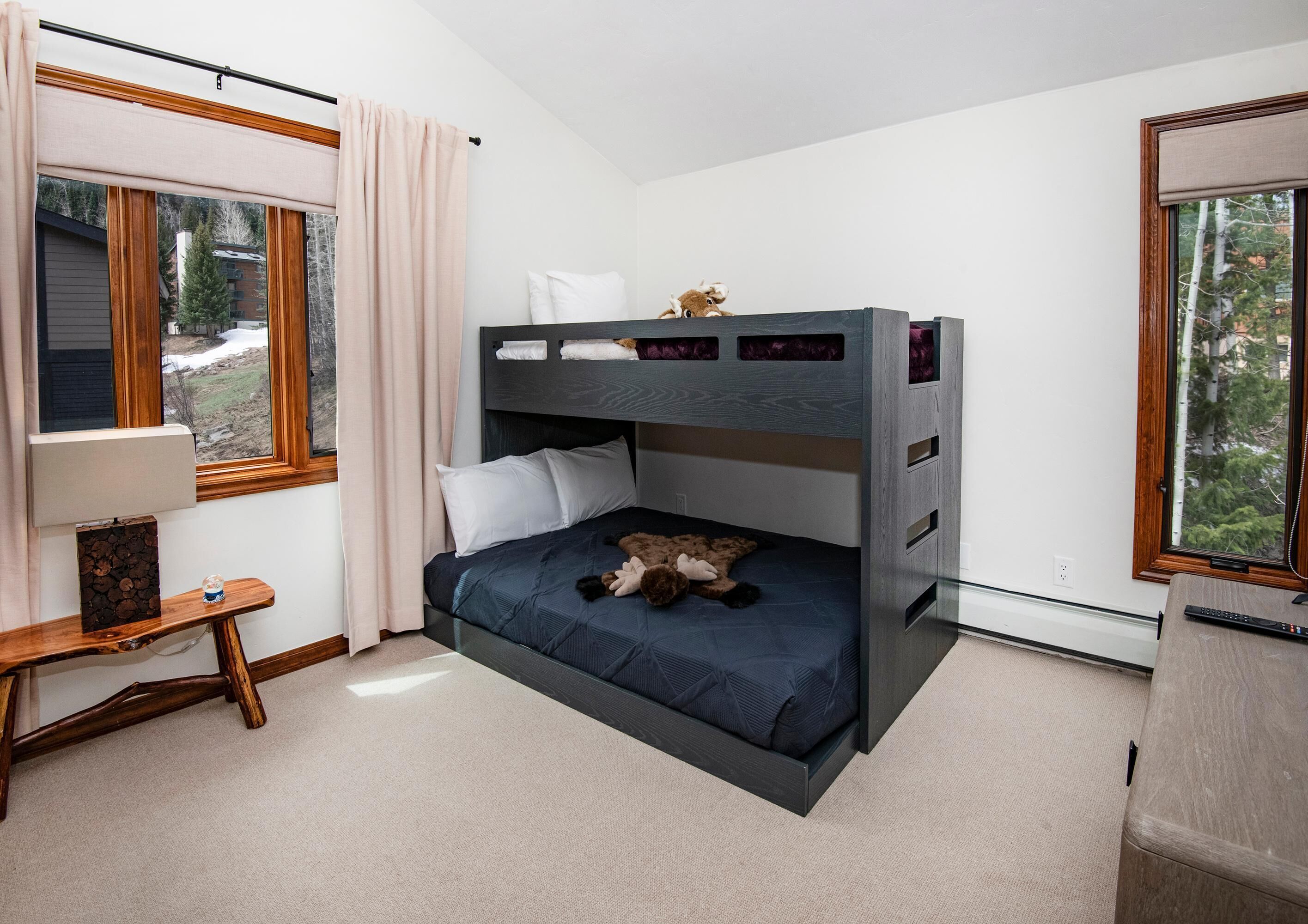 A bunk bed with a desk and chair, a window with curtains, and a view of trees outside.