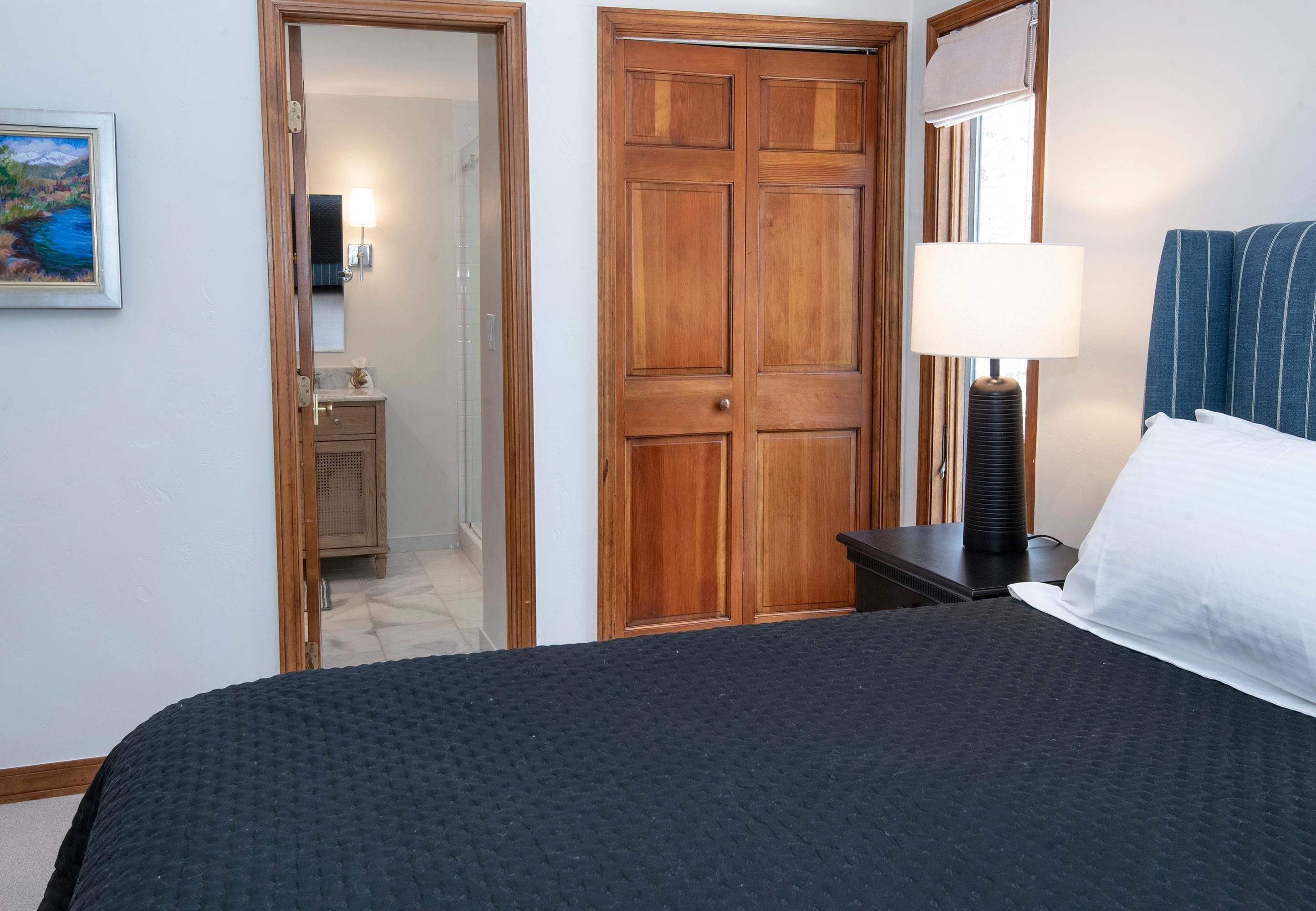 Een hotelkamer met een bed, een nachtkastje met een lamp, een houten garderobe en een badkamer die door een open deur zichtbaar is.