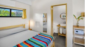 Hotellihuone, jossa on sänky, yöpöytä, peili, ikkuna verhoilla, kylpyhuone, jossa on pesuallas ja peili, sekä huonekasvi.