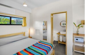 Habitación de hotel con cama, mesita de noche, espejo, ventana con cortinas, baño con lavamanos y espejo, y una planta.