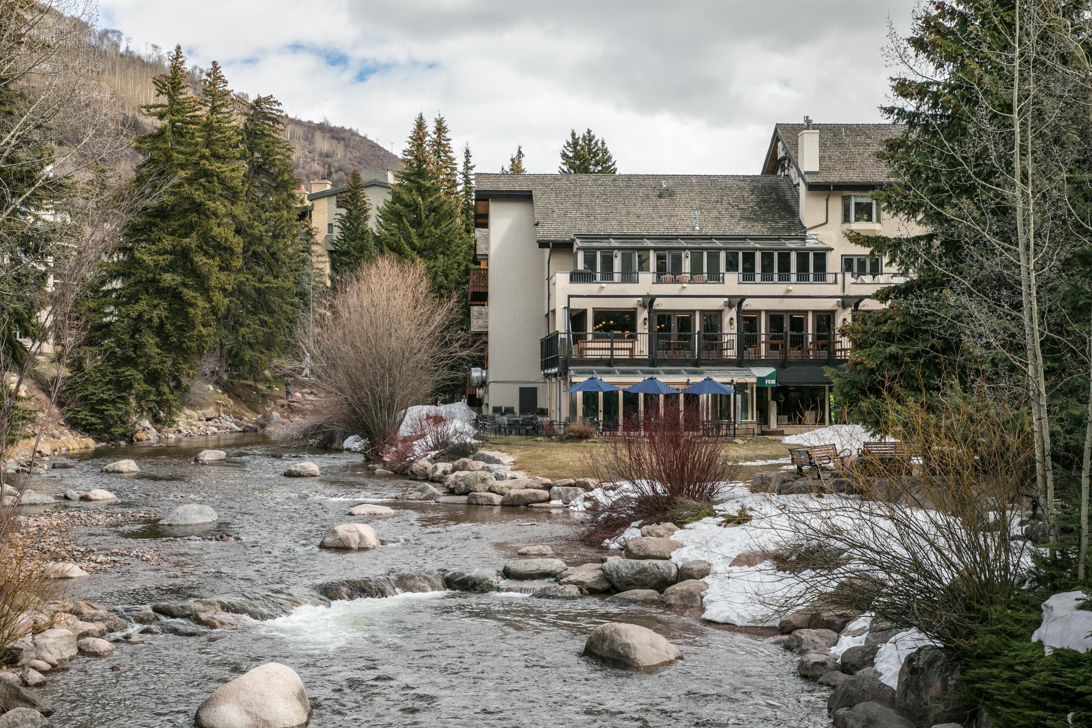 A riverside hotel with a stone facade, outdoor seating, and a mountain backdrop.