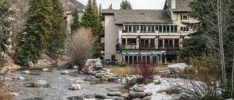 A riverside hotel with a stone facade, outdoor seating, and a mountain backdrop.