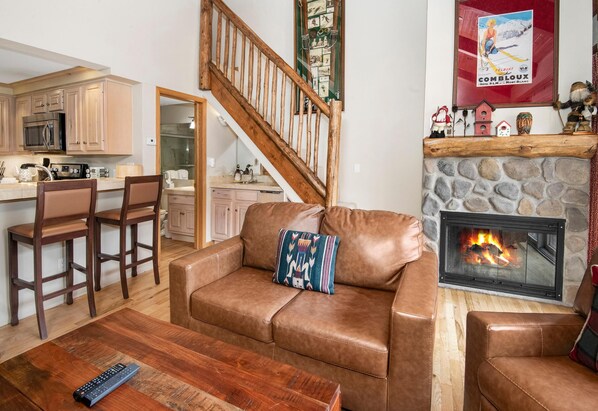Ein Wohnzimmer mit braunem Ledersofa, einem Couchtisch, einem Kamin und einer Küchenzeile mit Holzausbau und einer Mikrowelle.