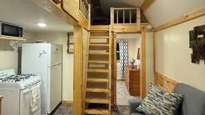 A compact kitchen with a gas stove, a white refrigerator, and a microwave on a shelf. A wooden staircase leads to a loft area with a window. - Cute, comfy, clean, safe, quiet, welcoming community in Madison. Pet-friendly (Madison)