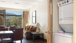 A room with a dining area, a sofa with cushions, a lamp, a view of the outdoors, and a washing machine. - Riverfront Resort & Spa 421a, 1BD Condo With Mtn Views (Avon)
