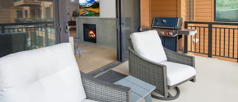 A balcony with wicker chairs and a grill, a fireplace with a fire, and a TV mounted on the wall.
