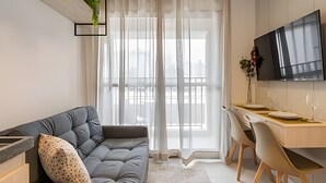 A modern living room with a sofa, a dining area with a table and chairs, a flat-screen TV mounted on the wall, and a large window with sheer curtains. - Aconchego Teodoro (São Paulo)