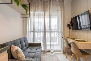 Una sala de estar moderna con un sofá, un comedor con mesa y sillas, un televisor de pantalla plana montado en la pared y un ventanal con cortinas ligeras.