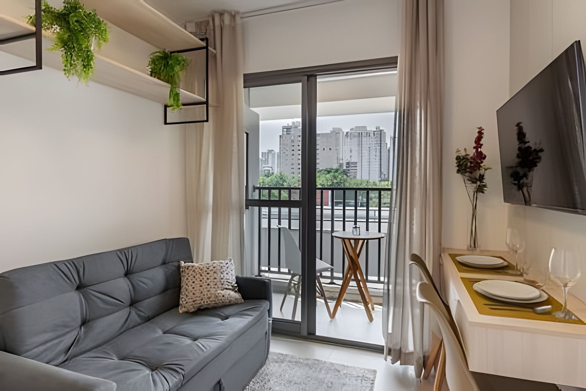 A modern apartment with a grey sofa, a dining area with a small round table, and a balcony with a city view.