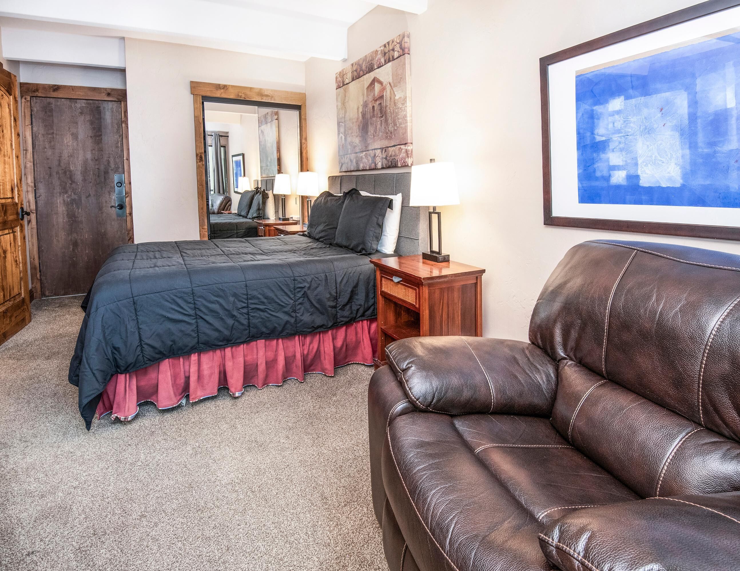 A hotel room with a bed, a leather sofa, a nightstand, a lamp, a mirror, and artwork on the walls.