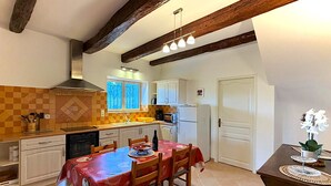 A kitchen with a dining table, chairs, a refrigerator, and a window with curtains. - "Le Colombier" 3★ Air-Conditioned Vacation Rental - Peace and Nature in the Heart of the Vineyards (Vinsobres)