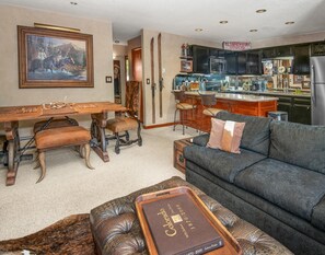 A dining area with a wooden table and chairs, a bar with stools, a kitchen with modern appliances, and a living room with a sofa and a book on a tray. - Scorpio 101, 2BD Vail Condo With Wood Fireplace (Vail)