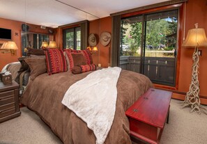 A bedroom with a large bed, bedside lamps, a wooden nightstand, a red bench, and a view of the outdoors through a sliding door. - Scorpio 101, 2BD Vail Condo With Wood Fireplace (Vail)