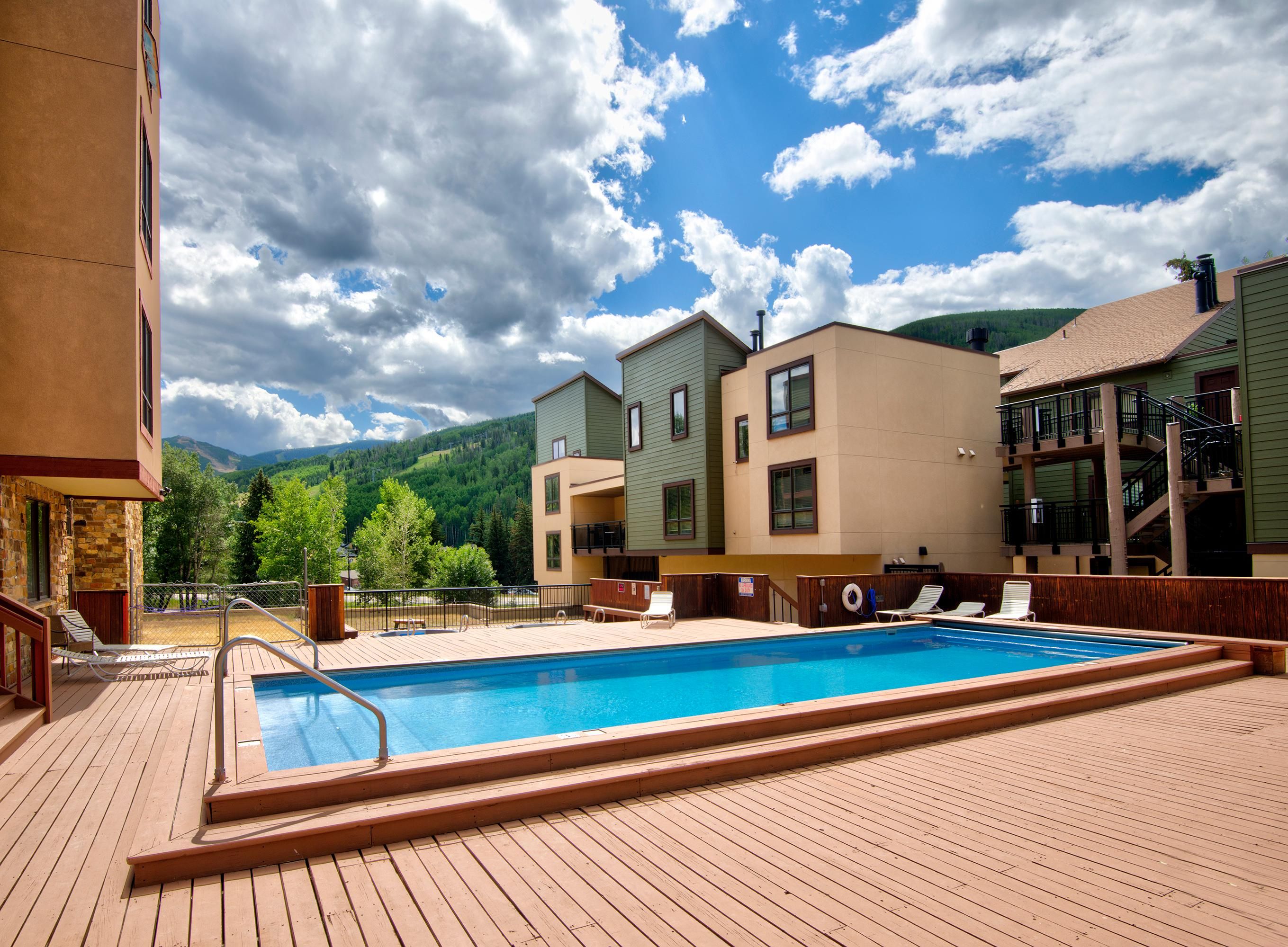 A pool area with a wooden deck, lounge chairs, and a view of mountains.