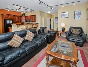 Ein Wohnzimmer mit einer schwarzen Ledersofa, einem Glastisch und einem Ledersessel. Es gibt eine Küchenzeile mit Holzschränken und einer Mikrowelle.