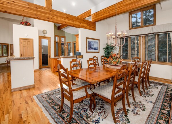 A spacious dining area with a large wooden table and chairs, a chandelier, and a view of trees through large windows. - 4BD Private Arrowhead Home on Golf Course (Edwards)