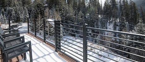 A snow-covered deck with a railing overlooking a forested mountain area.