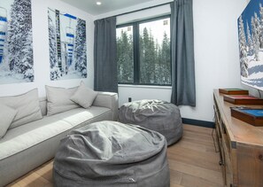 Ein modernes Wohnzimmer mit einer Couch, zwei Sitzsäcken, einem Holztisch mit Büchern und einem Fenster mit Blick auf schneebedeckte Bäume.