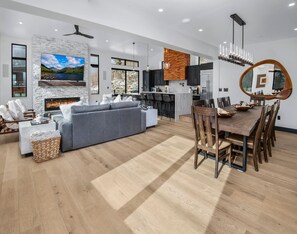 A modern living room with a fireplace, a dining area with a wooden table and chairs, and a kitchen with dark cabinets and a wooden accent wall. - Big Horn House, 4387 Columbine (Vail)