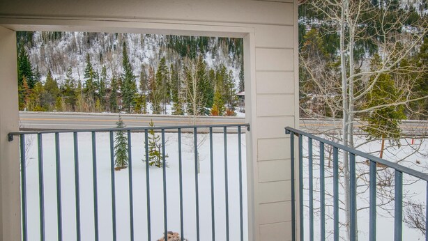 A covered porch with a metal railing, a view of a snowy landscape with trees, and a road in the background. - Spruce Way 209, 2BD East Vail Condo (Vail)
