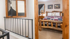 A bedroom with a rustic wooden bed frame, a large window with blinds, a ceiling fan, and two framed pictures on the wall. - Spruce Way 209, 2BD East Vail Condo (Vail)