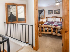 A bedroom with a rustic wooden bed frame, a large window with blinds, a ceiling fan, and two framed pictures on the wall. - Spruce Way 209, 2BD East Vail Condo (Vail)