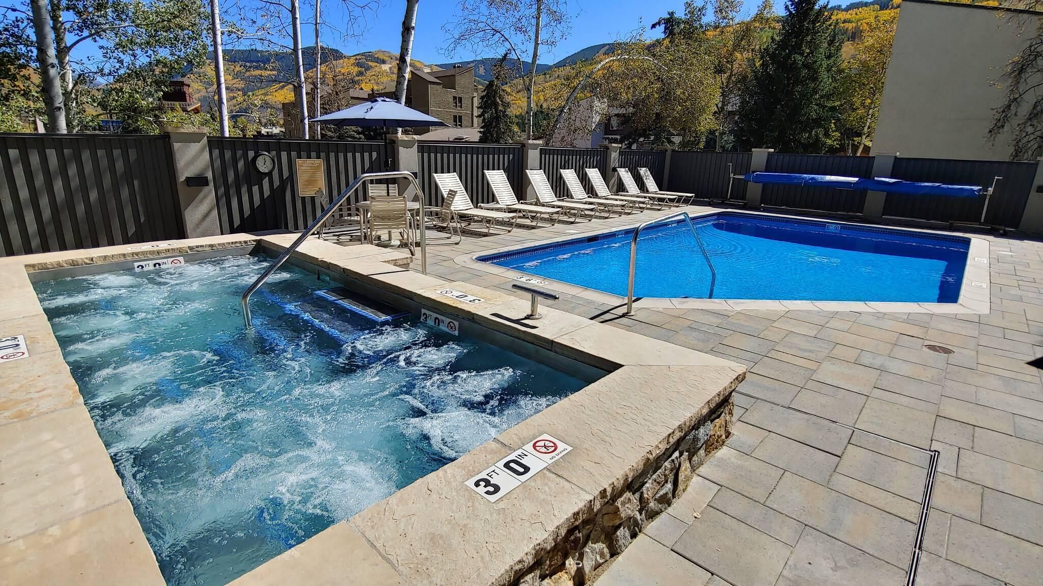 A hot tub and a pool with lounge chairs, a wooden fence, and a mountainous backdrop.