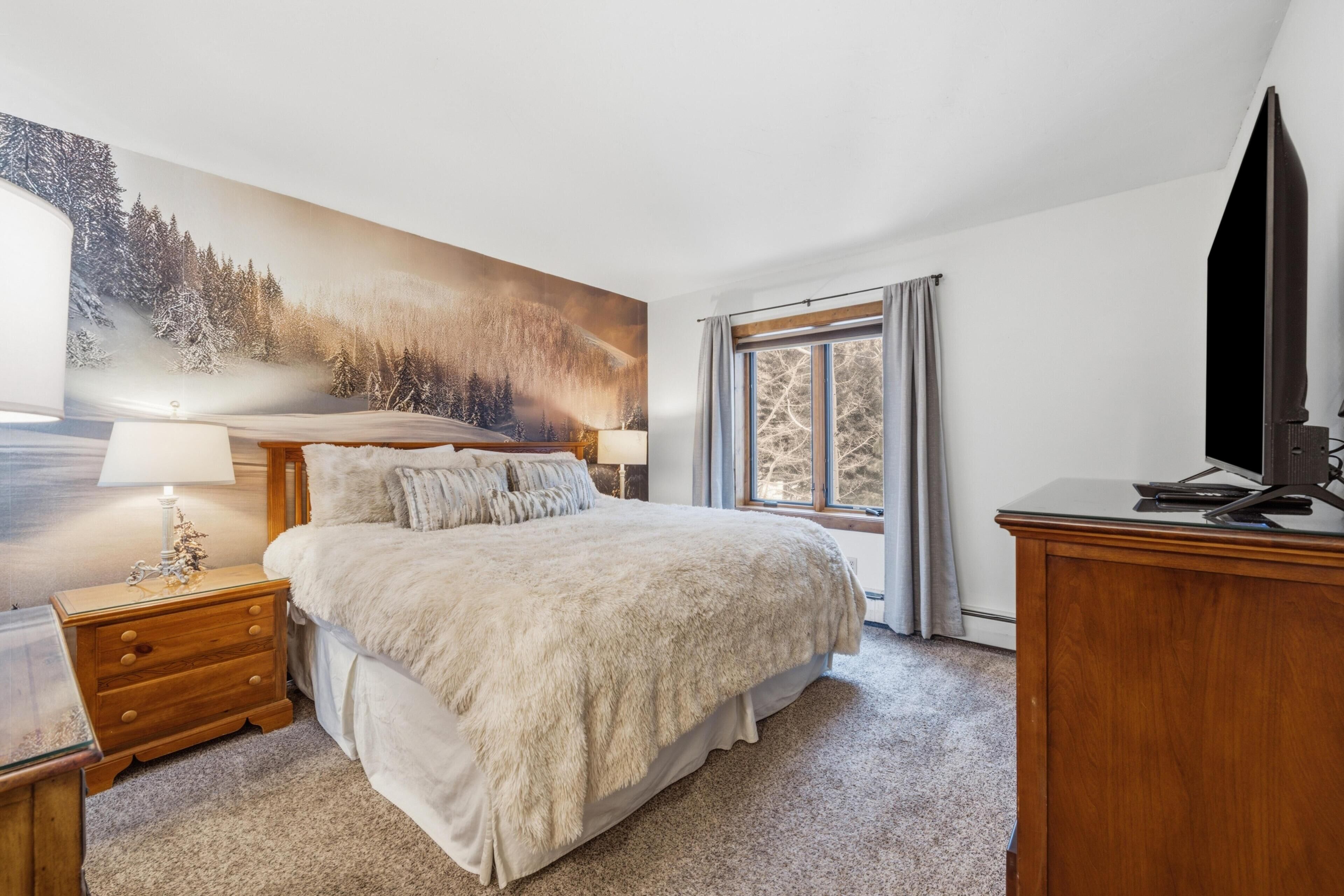 Een slaapkamer met een groot bed, een nachtkastje met een lamp, een houten dressoir, een televisie en een raam met gordijnen.