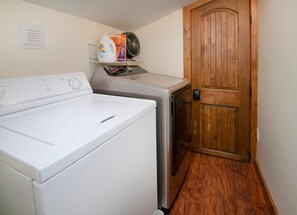 A laundry room with a front-loading washer and dryer, a wooden door, and a shelf with laundry supplies. - Timber Falls 1005, 3BD East Vail Condo With Amenities (Vail)