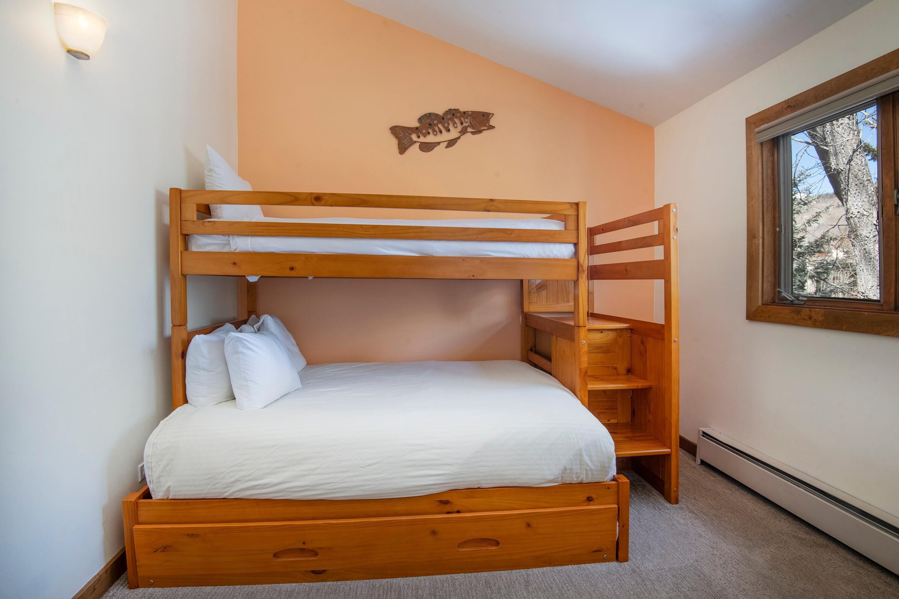 Habitación con literas, cabecero de madera, ropa de cama blanca, un adorno de pez en la pared y ventana con vista a árboles.