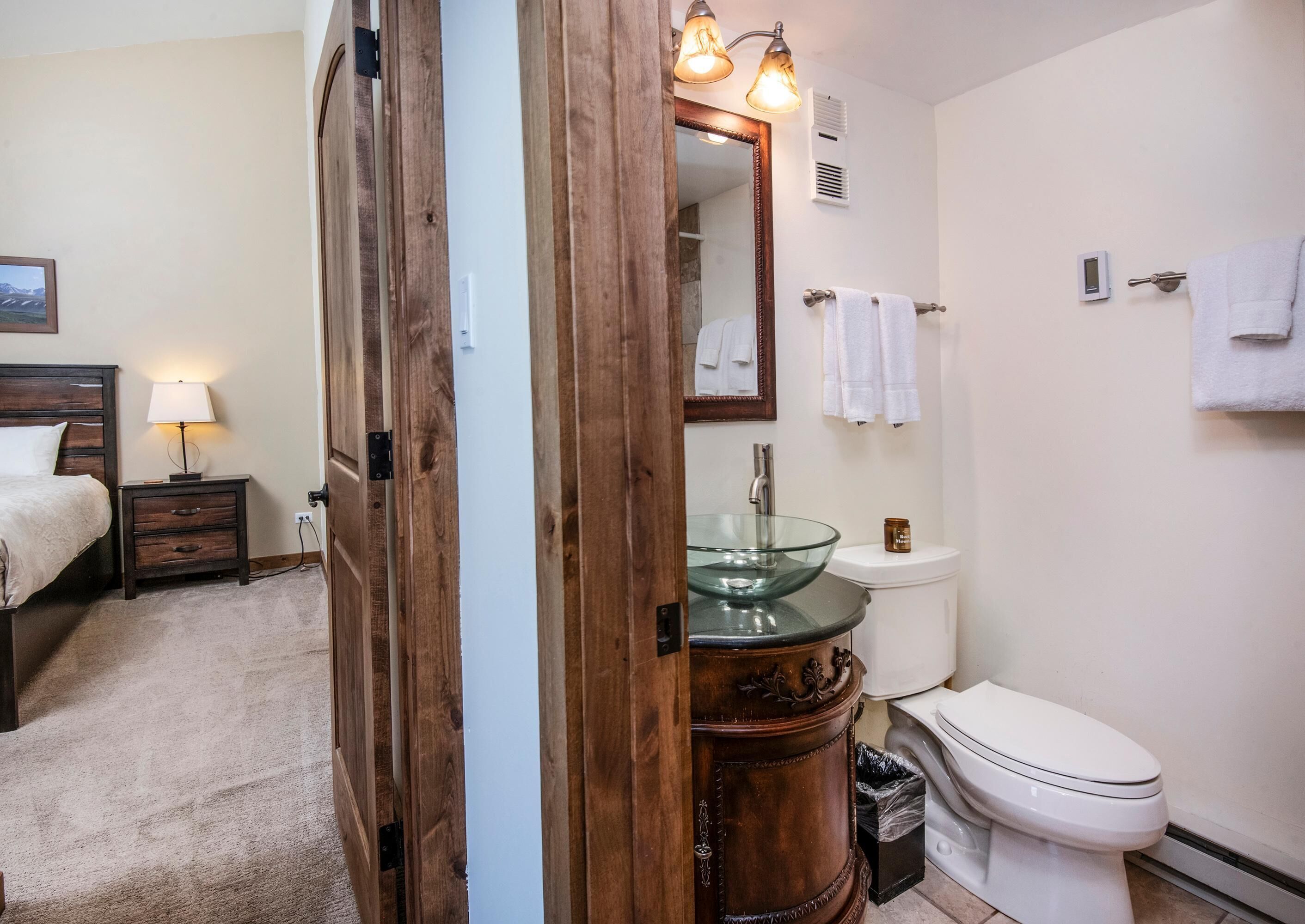 A bathroom with a glass bowl sink, a toilet, a wooden vanity, a mirror, and a bedside table with a lamp.