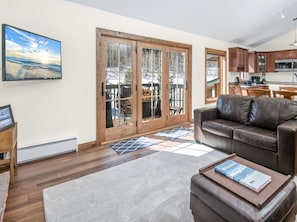 A living room with a leather sofa, a coffee table with a book, a TV on the wall, and a sliding glass door leading to a deck. - Timber Falls 1005, 3BD East Vail Condo With Amenities (Vail)