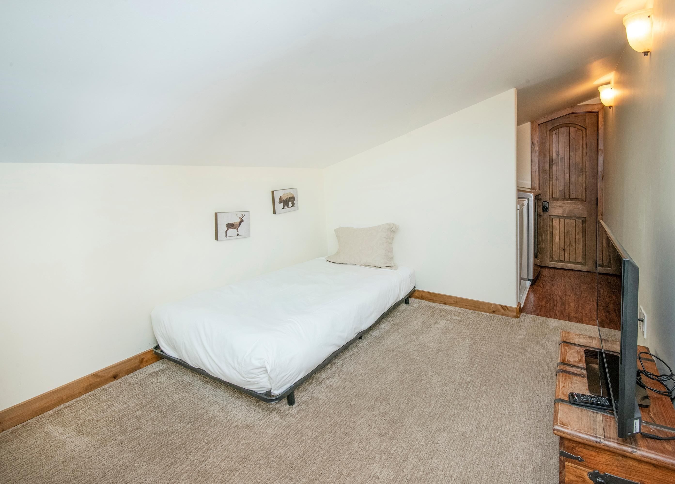 Una habitación pequeña con una cama individual, una puerta de madera, un escritorio pequeño con una computadora y una lámpara fijada a la pared.