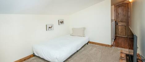 A small, single-bed room with a wooden door, a small desk with a computer, and a wall-mounted light fixture.