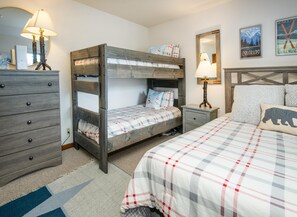 Ein Etagenbett mit Holz-Kopfteil, ein Nachttisch mit Lampe, eine Kommode und ein Bett mit karierter Decke.