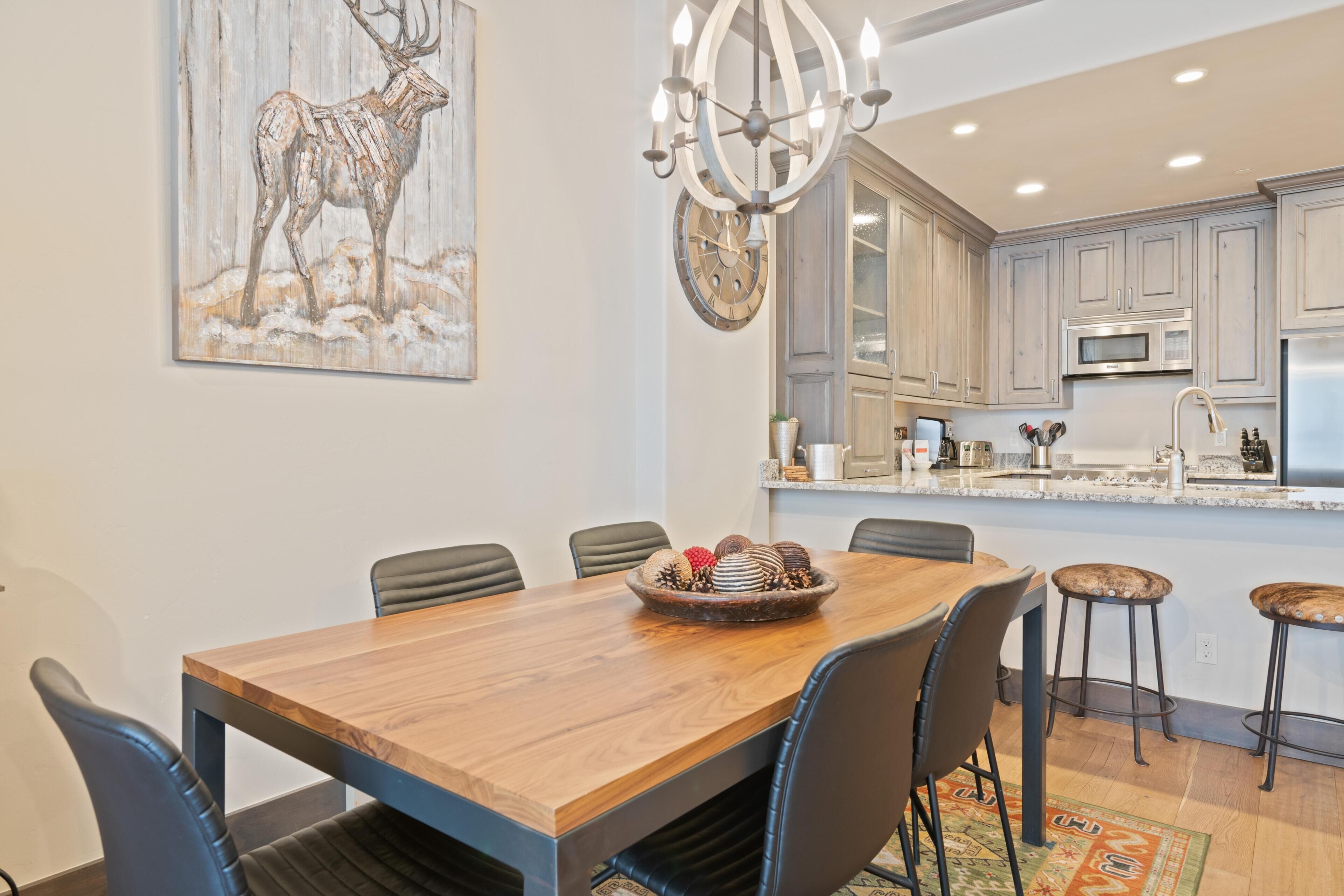 A dining area with a wooden table, chairs, a chandelier, and a kitchen in the background.