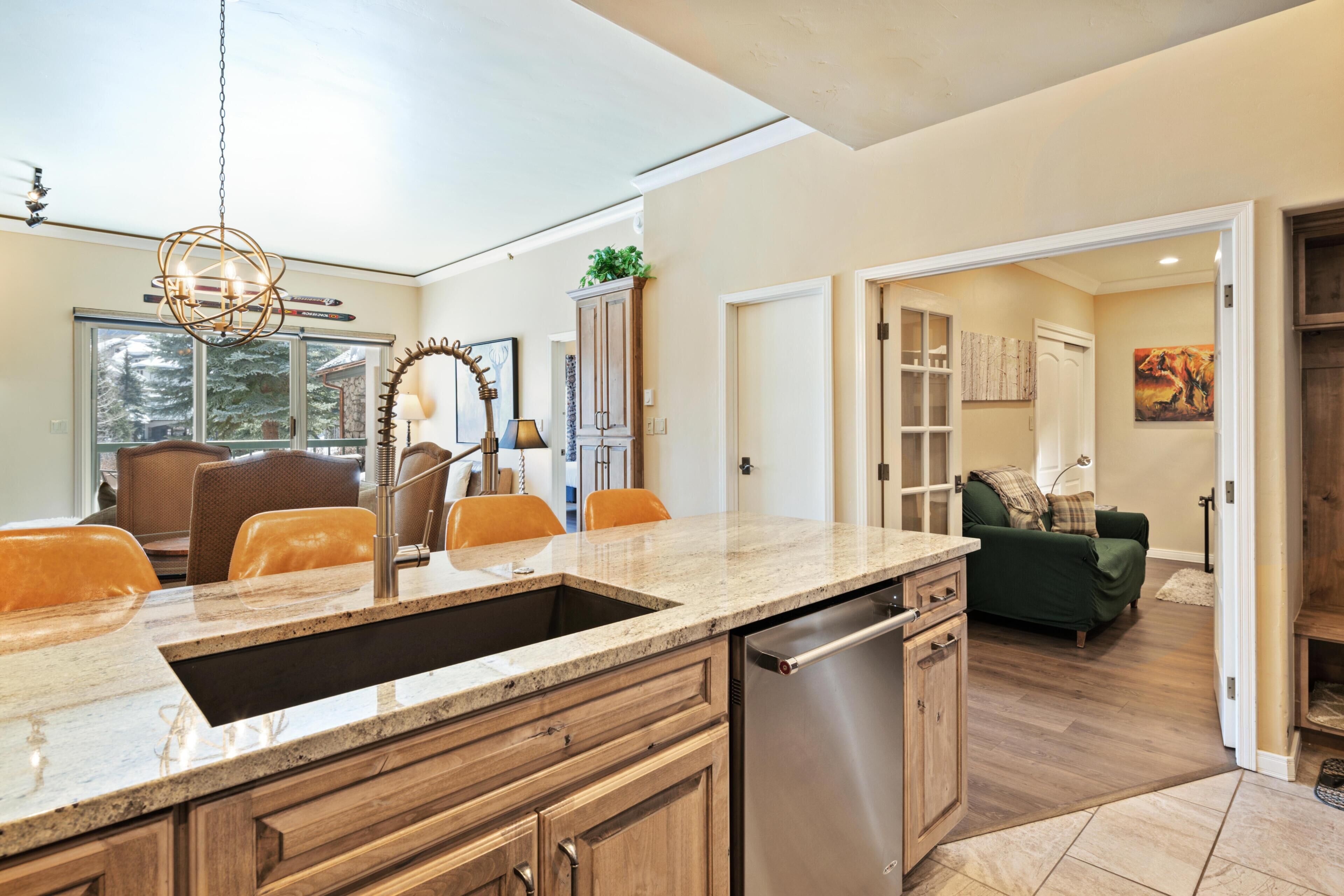 A modern kitchen with a granite countertop, stainless steel dishwasher, wooden cabinets, and a pendant light.