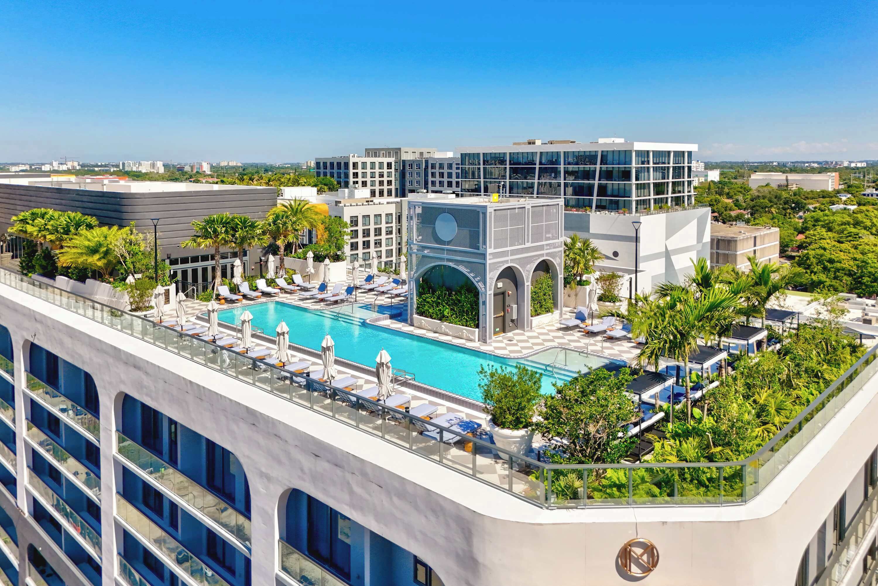 A rooftop pool with lounge chairs and umbrellas, surrounded by modern buildings and greenery.