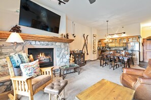Uma sala de estar com lareira de pedra, móveis de madeira, televisão montada acima da lareira e uma área de cozinha com armários de madeira e geladeira.