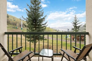 A balcony with two chairs and a small table, overlooking a grassy area with trees and a distant mountain range. - Borders Lodge L109 2BD Beaver Creek Ski-in, Ski-out Condo (Avon)
