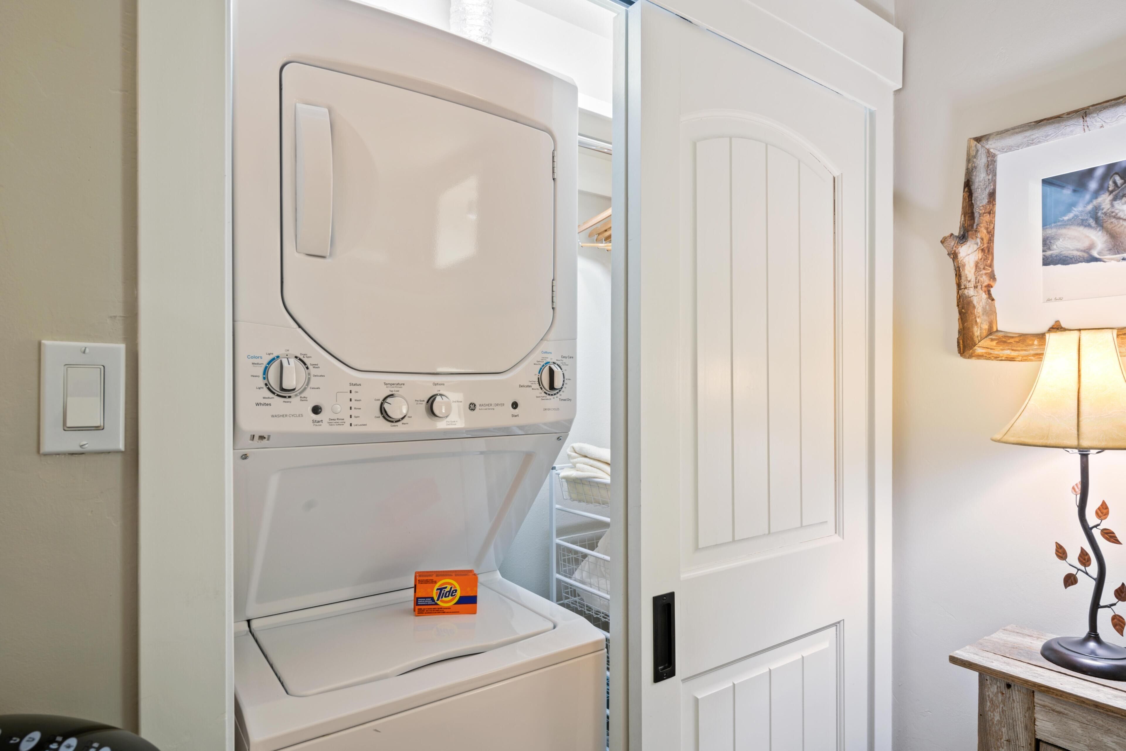 Una lavadora blanca con panel de control, una caja de detergente Tide y una mesa de madera con una lámpara.