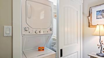 A white washing machine with a control panel, a Tide detergent box, and a wooden table with a lamp.