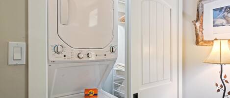 Una lavadora blanca con panel de control, una caja de detergente Tide y una mesa de madera con una lámpara.