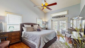 Une chambre à coucher comprenant un lit, une commode, une chaise, un ventilateur de plafond et de grandes fenêtres.