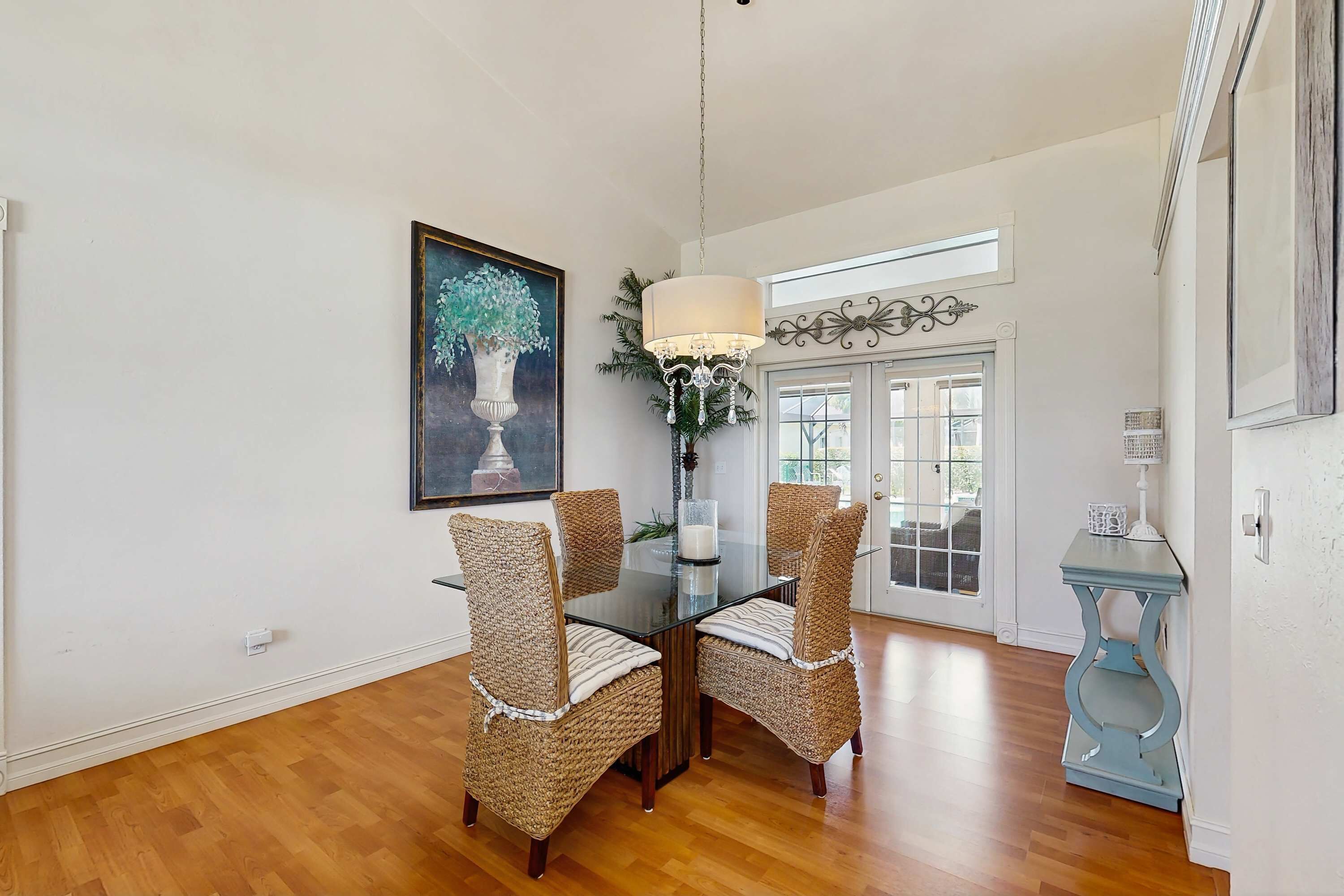 A dining area with wicker chairs, a glass table, a chandelier, and a painting on the wall.
